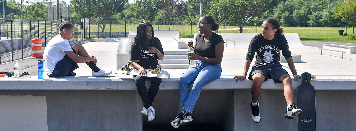 A group of friends sitting on the ledge of a skate park holding boards.