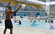 an instructor on the indoor pool deck leads a class in a series of exercises which they perform in a pool filled with water