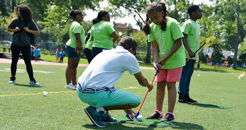 An instructor teaches a camper how to play golf on the turf in the park.