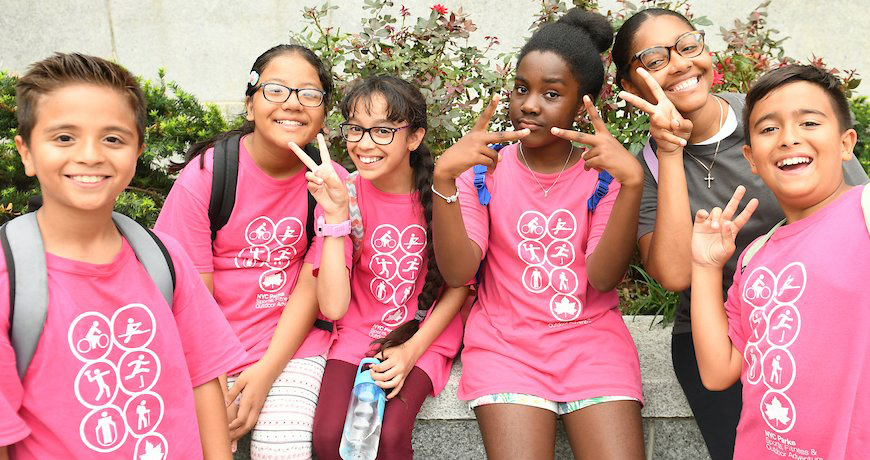 A group of summer camp kids in their pink Summer Camp t-shirts poses for a photo in the park.