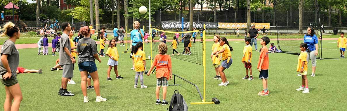 Children in uniform playing volleyball together while coaches look on.