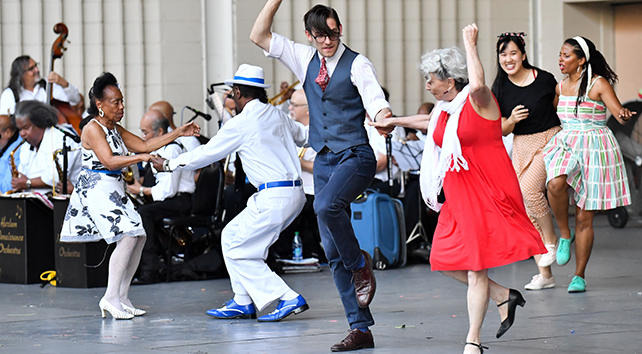 Dance partners in retro outfits swing while a jazz orchestra plays.