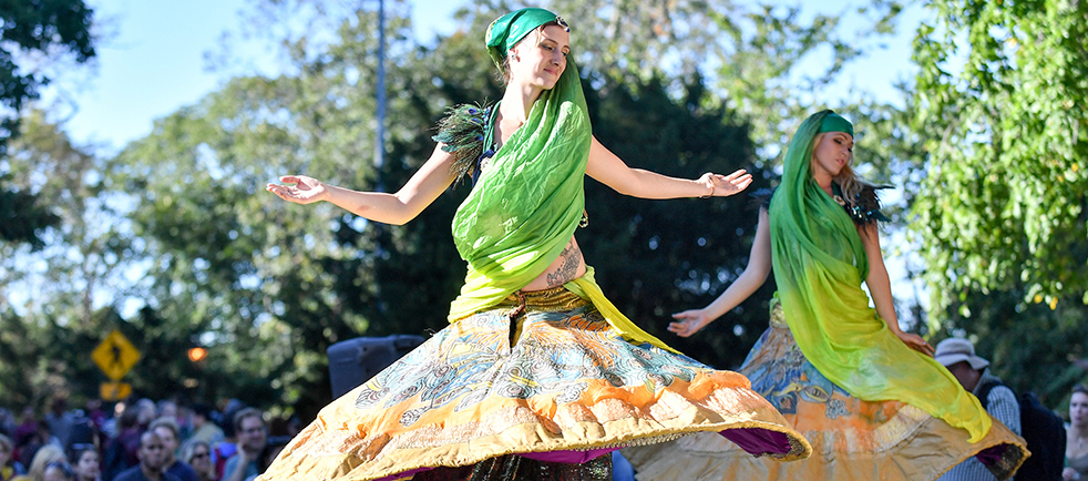 A pair of women in a park are wearing ornate sashed dresses and moving in rhythm.