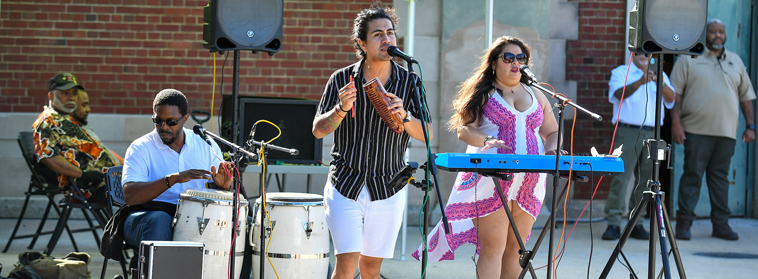 A band plays Latin percussion instruments, including a guiro and conga drums, in front of a recreation center.