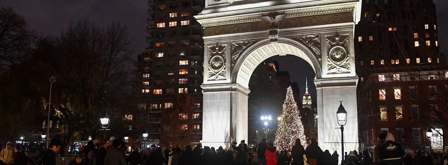 A Christmas tree is lit up at night under the famous Washington Square Arch monument while a crowd gathers around.