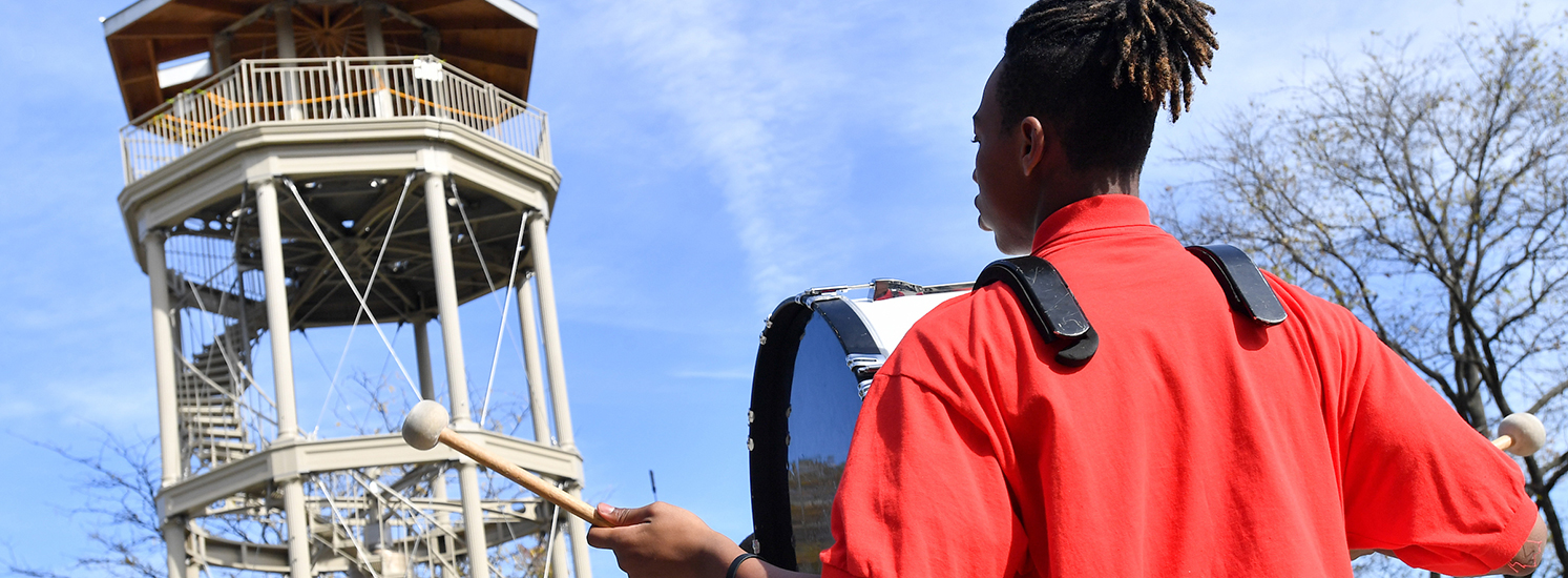 A drummer in a marching band looks at a historic spiral tower during an event.