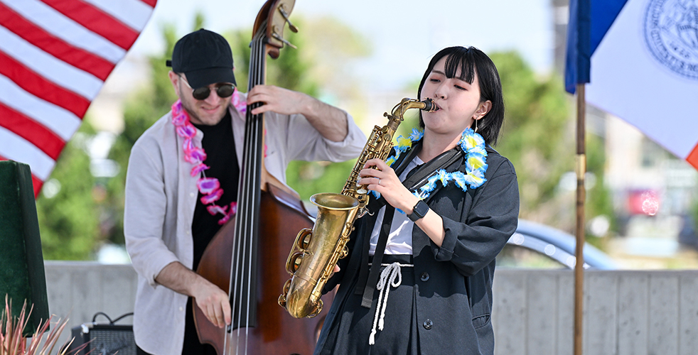 On a summer day, two musicians can be seen. A woman leans back in performance playing the saxophone, while a man behind her plays bass.