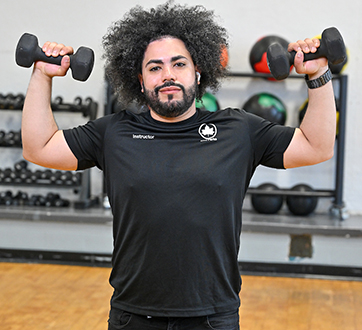 A young fit man holds two small dumbbells up on either side of his body, using proper form. His t-shirt has the NYC Parks logo and reads 