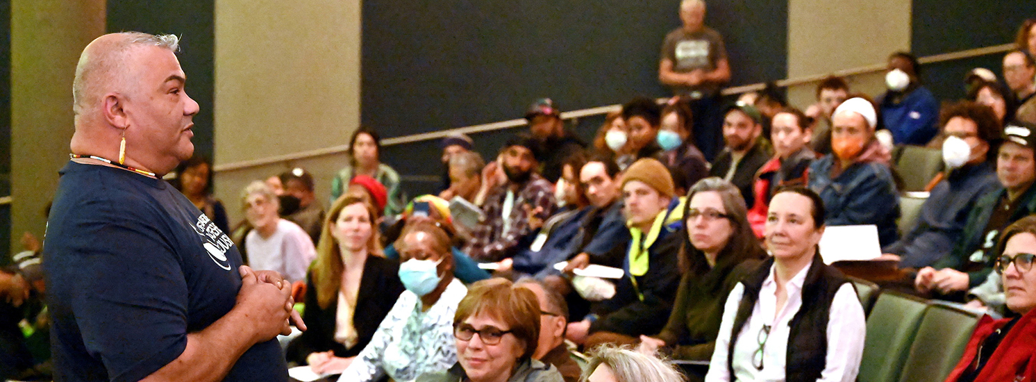 A man is standing in an auditorium, speaking to a seated crowd.