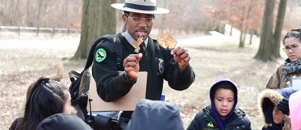 A person in a ranger uniform is holding up two leaves and explaining the differences to a group of children.