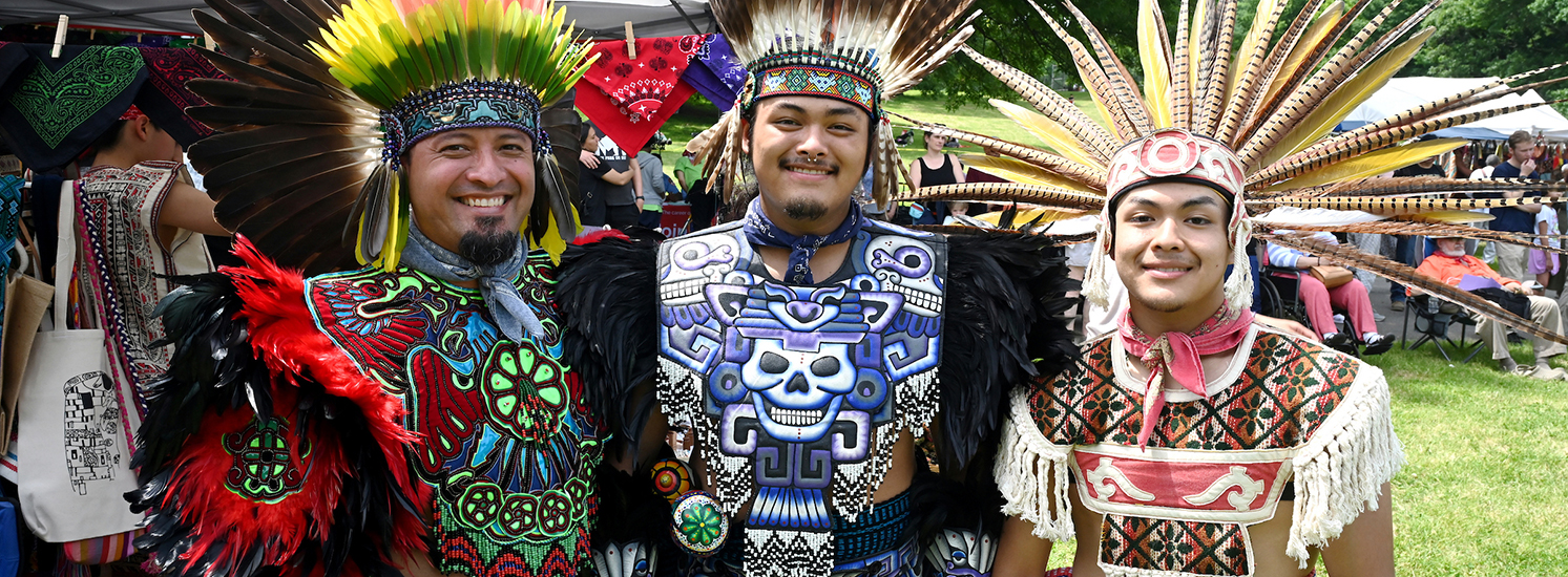 Three young men smile at the camera while dressed in colorful ceremonial outfits, with intricate designs and large headdresses.