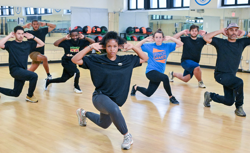 A group of people in formation in a room, lunging with one leg crossed over another, holding hands behind their head. A young woman wearing a black t-shirt with an NYC Parks logo is in front of the group, looking at the camera.